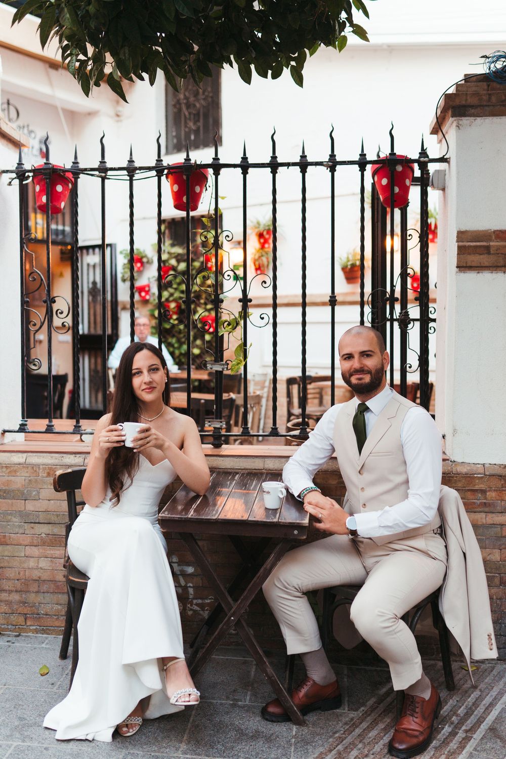 A couple in wedding attire sits at a small table near a decorative iron gate with red flowers.