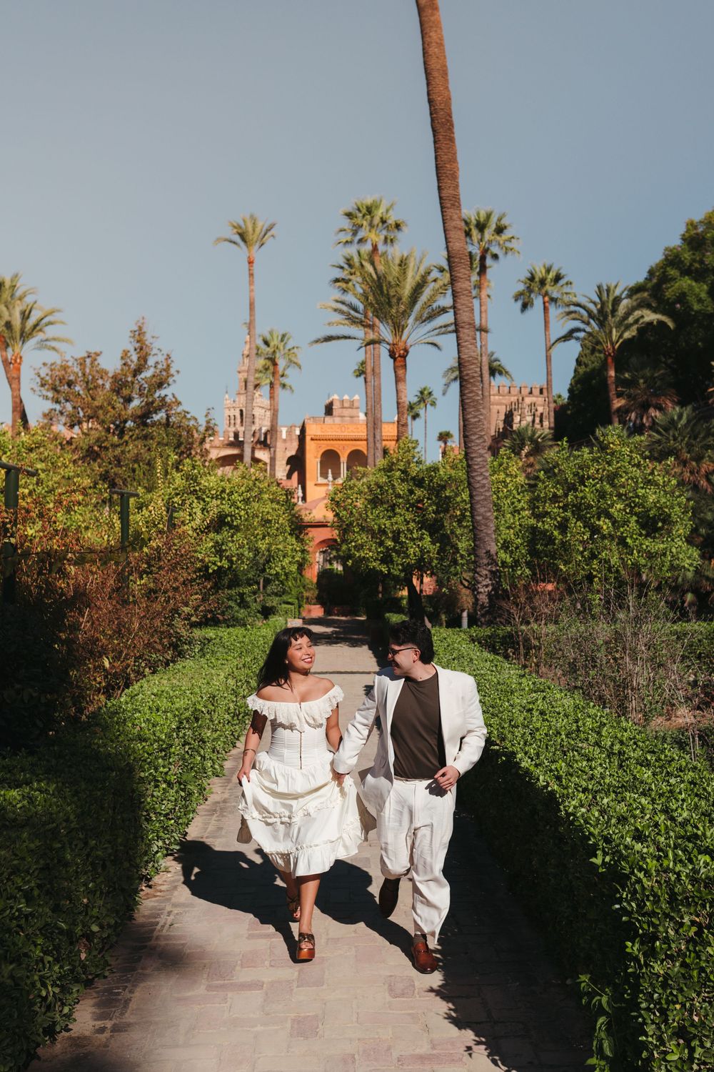 A couple strolls down a palm-lined path towards a Moorish-style building.