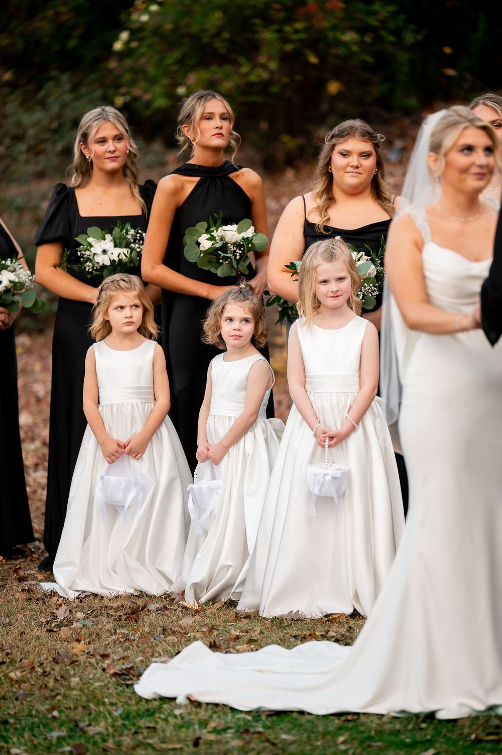 Row of flower girls in white dresses stand together during outdoor wedding ceremony.
