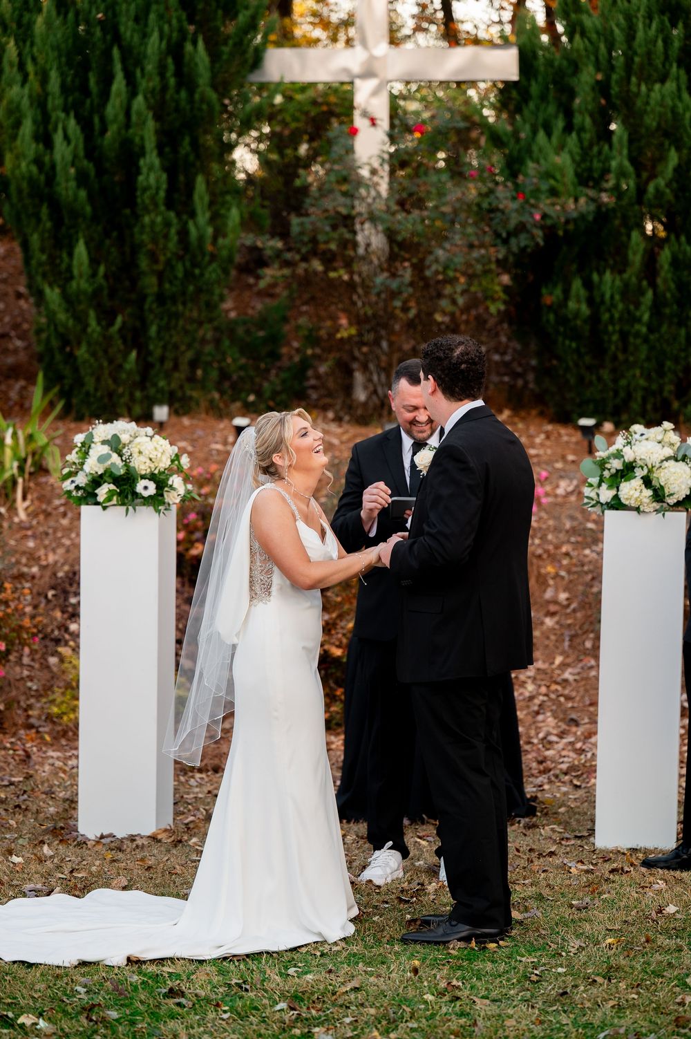 Wedding ceremony moment captured outdoors with white pillar decorations and evergreen backdrop.