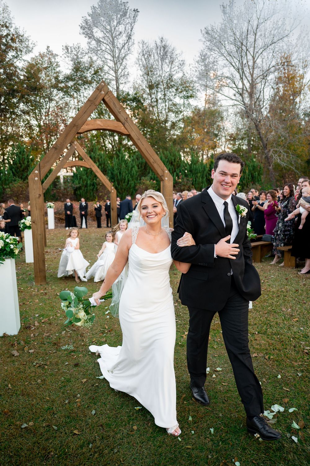 Joyful wedding couple celebrates under wooden arch during outdoor autumn ceremony.