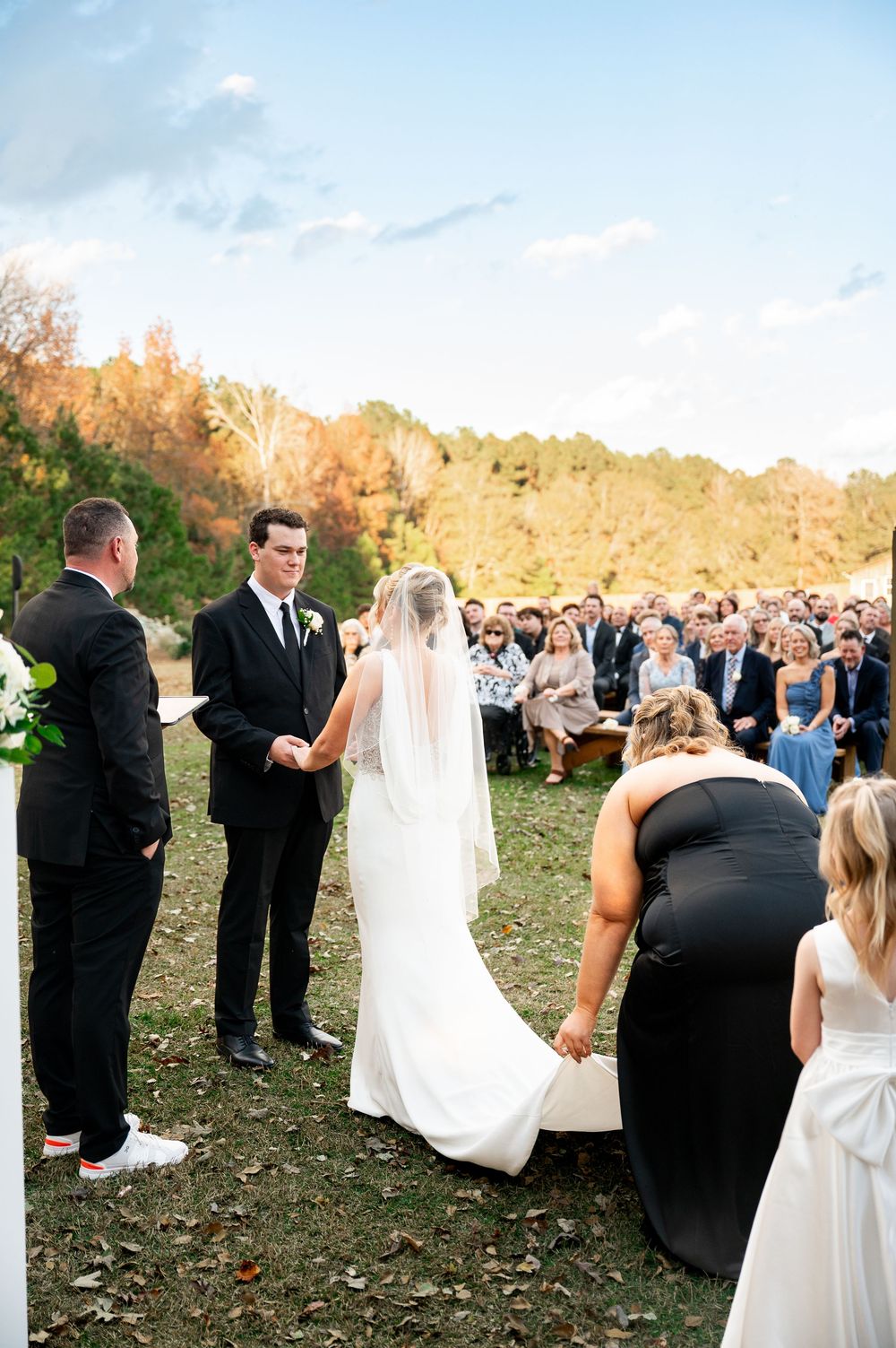 Outdoor autumn wedding ceremony with guests gathered on grassy hillside during golden hour.