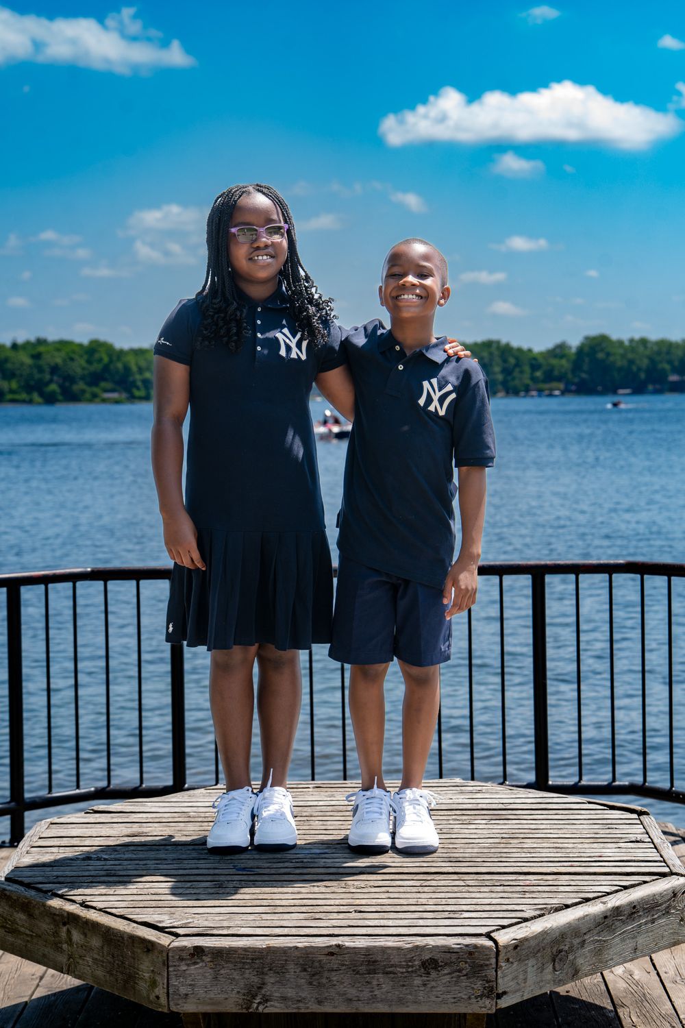 Two people wearing matching NY Yankees outfits stand on a wooden dock by a serene lake on a sunny day.