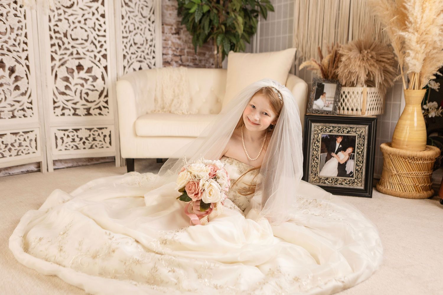 A bride in an elegant lace wedding gown sits gracefully in a cream armchair surrounded by decorative screens and pampas grass.