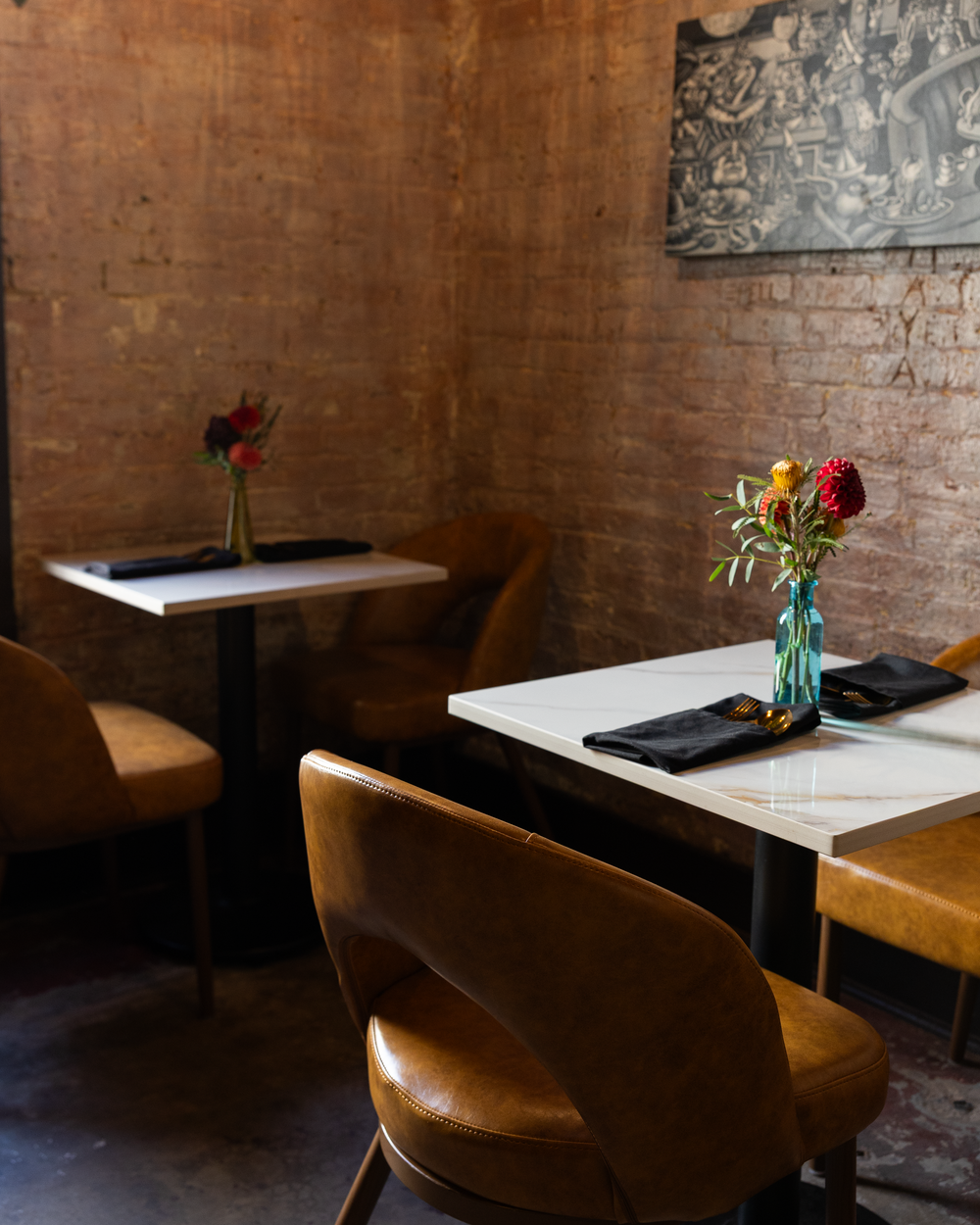 Warm restaurant dining area featuring exposed brick walls, leather seating, white marble tables with floral accents.