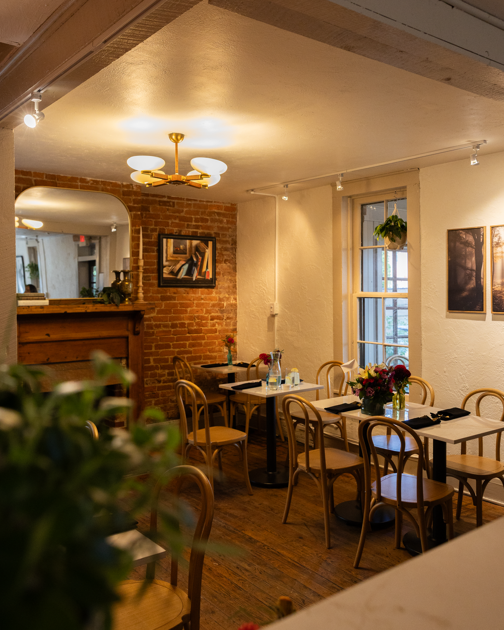 Charming restaurant interior with exposed brick, wooden chairs, white walls, artwork, and warm ambient lighting.