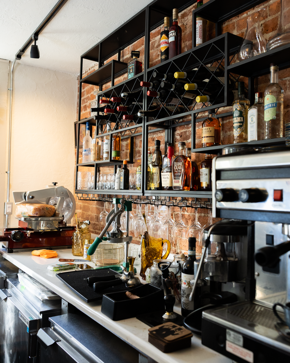 Restaurant bar shelf with wine bottles, liquor, glassware, and espresso machine against exposed brick wall.