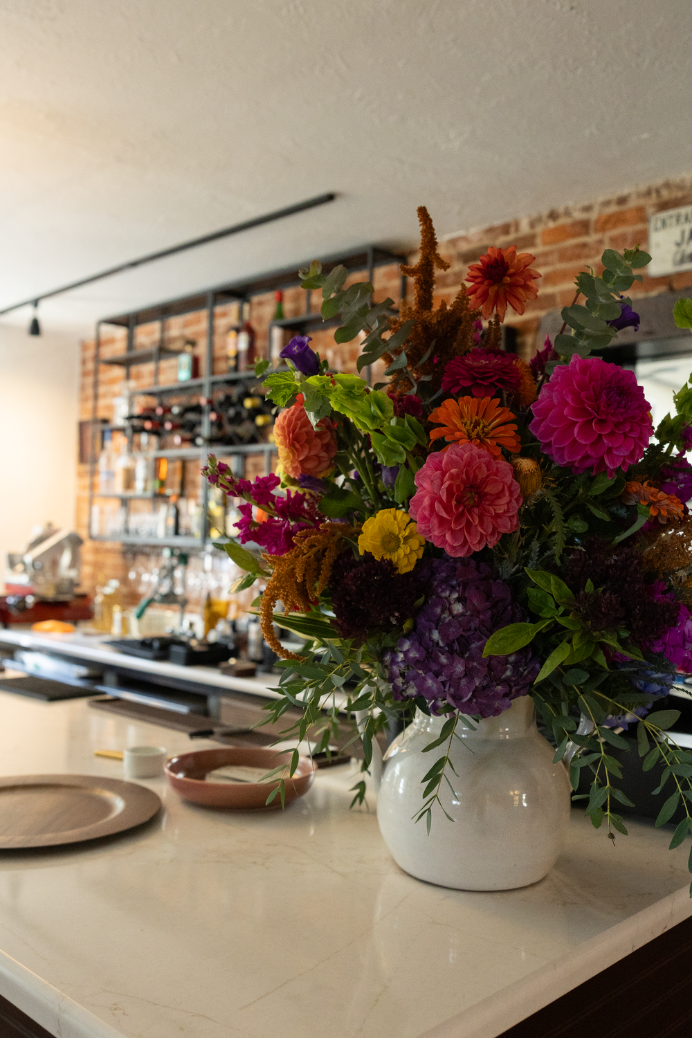 Colorful floral arrangement with dahlias and greenery in white vase on marble counter in restaurant.