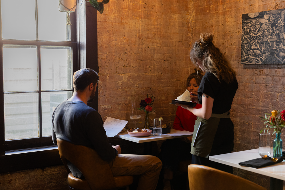 Server taking order from seated customer in cozy brick-walled restaurant dining room.