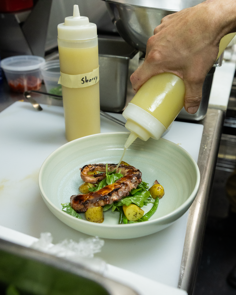Chef plating grilled meat dish with roasted vegetables in green bowl, drizzling sauce from squeeze bottle.