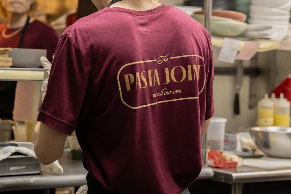 Staff member wearing maroon The Pasta Joint restaurant t-shirt standing in kitchen.