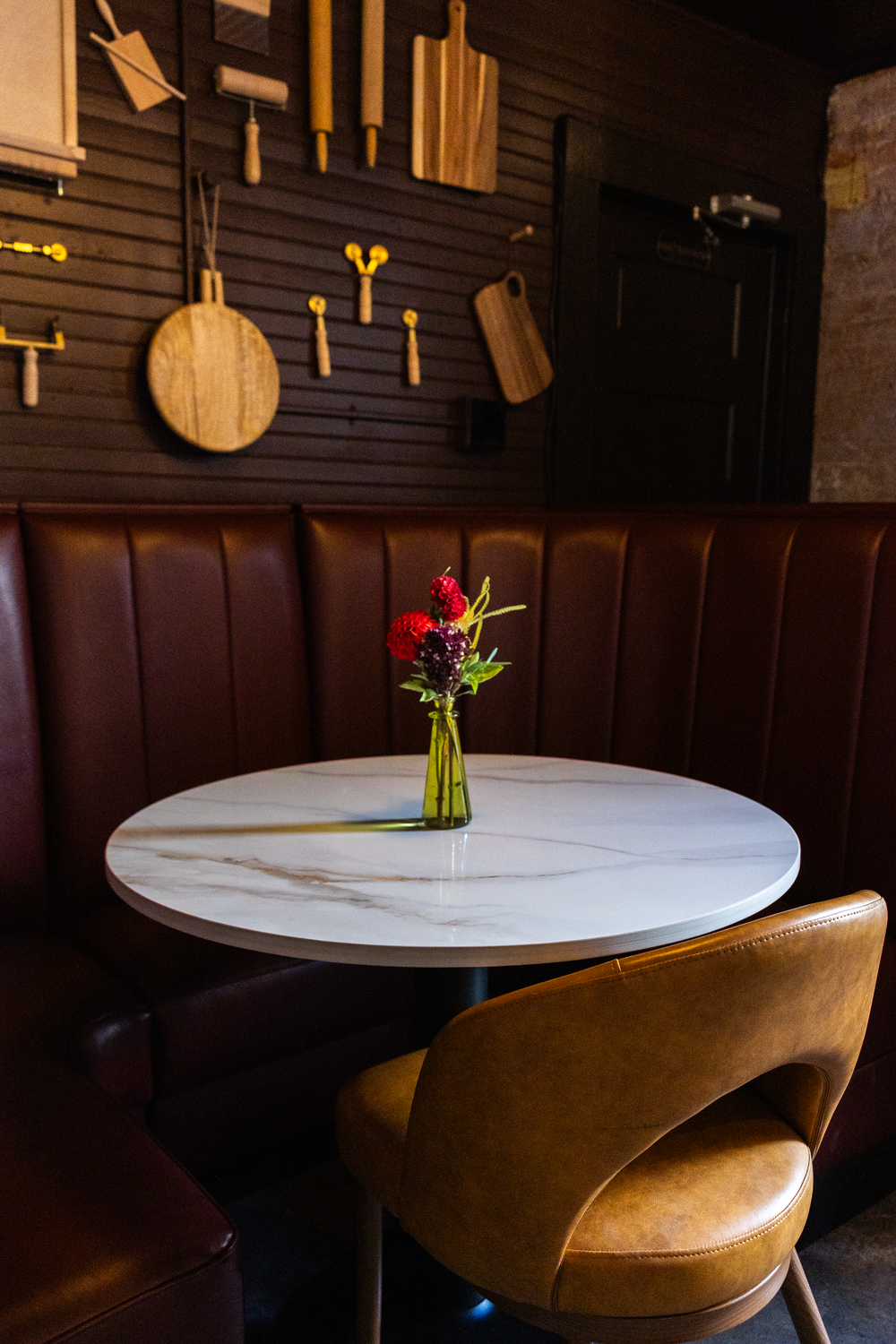 Intimate restaurant booth with white marble round table, red flower centerpiece, and wooden kitchen decor wall display.