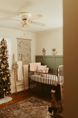 Elegant sage green nursery with iron crib, Christmas tree, and botanical wall art for a baby girl.