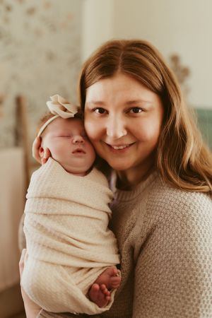 Smiling mother holding her sleeping swaddled newborn baby girl with a bow headband against her cheek.