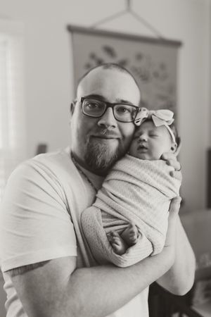 Black and white portrait of a bearded father holding his swaddled newborn baby girl with a bow.