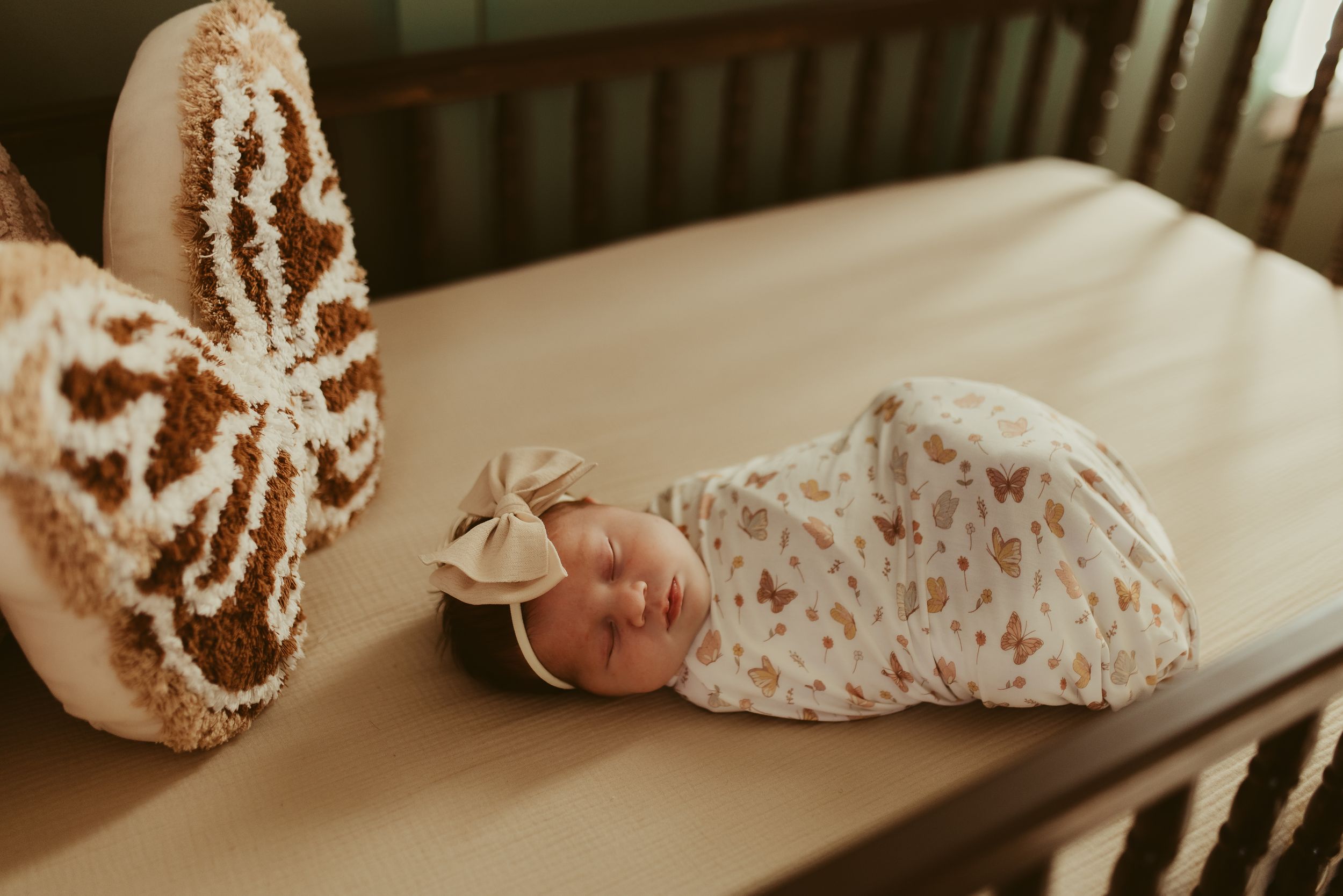 Sleeping newborn girl in butterfly swaddle with bow headband lying in wooden crib beside plush toy.