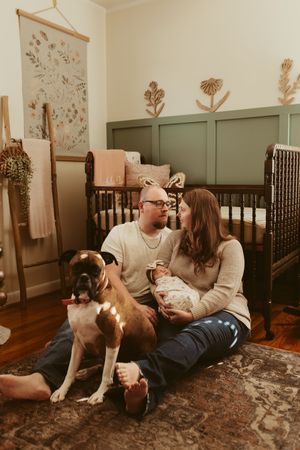New parents sitting on nursery floor with boxer dog holding newborn in beautifully decorated baby room.