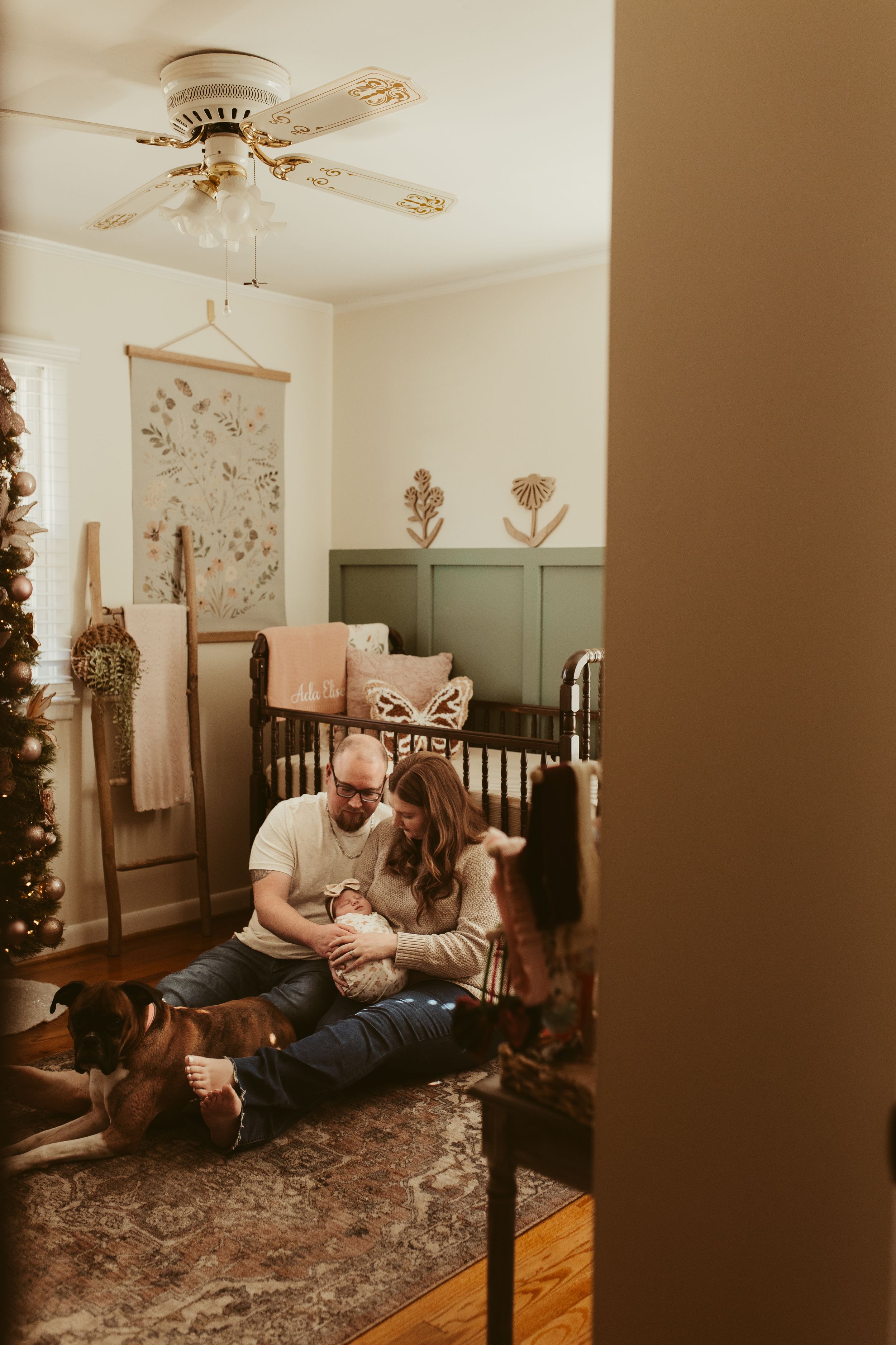 Young family with newborn and boxer dog sitting in green and neutral toned nursery near Christmas tree.