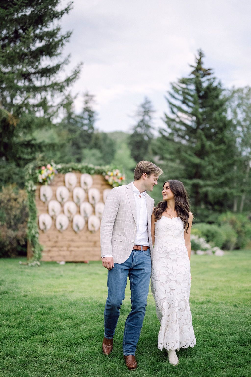 A romantic couple walks hand in hand on a green lawn with evergreen trees and rustic decor in the background.