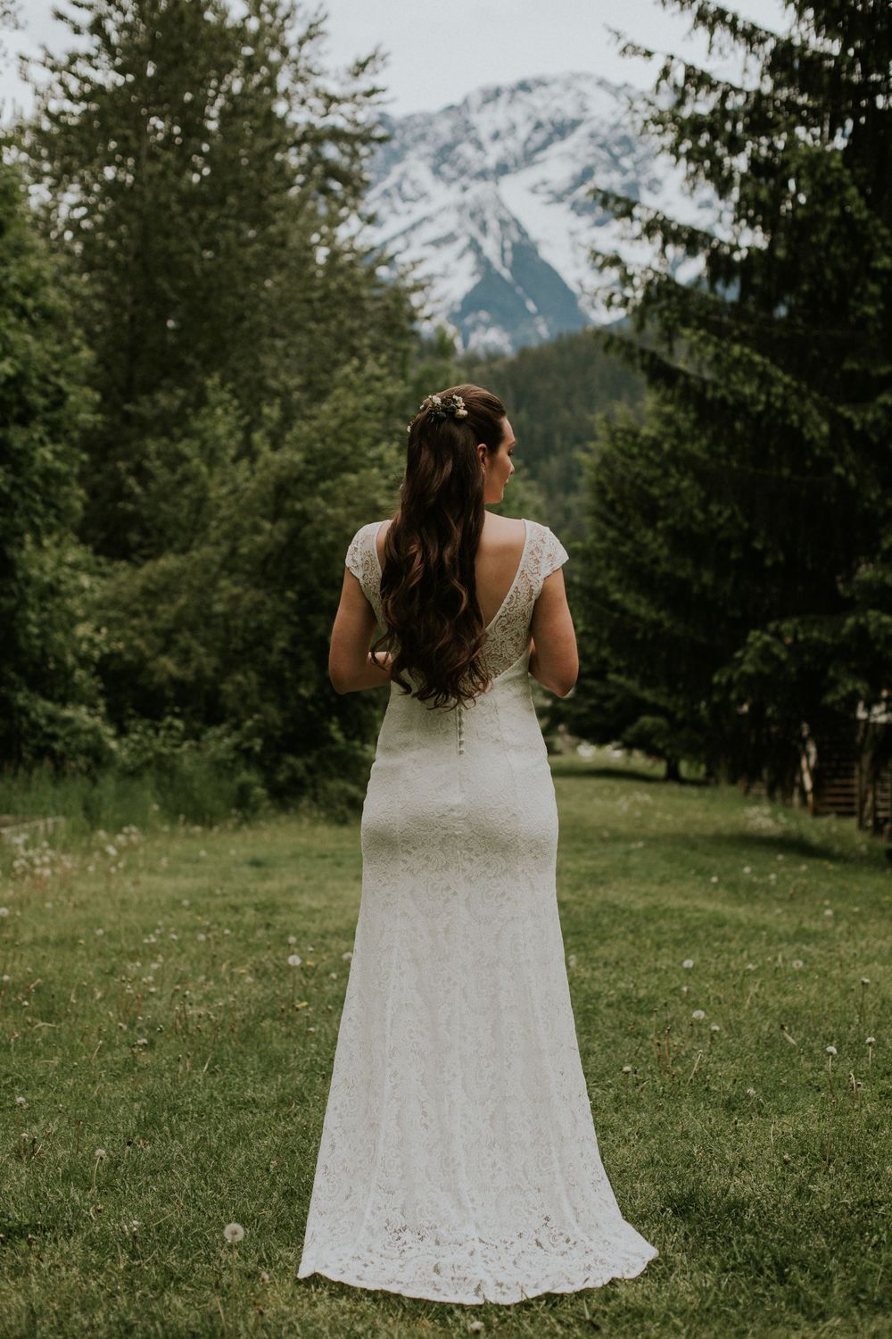 A bride in a fitted lace gown looks out at snow-capped mountains during an outdoor wedding.
