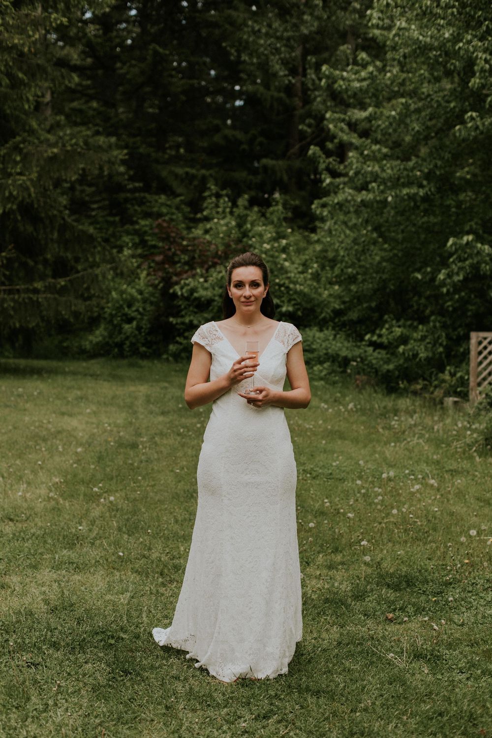 A person in a white lace dress stands in a grassy outdoor setting surrounded by trees and greenery.