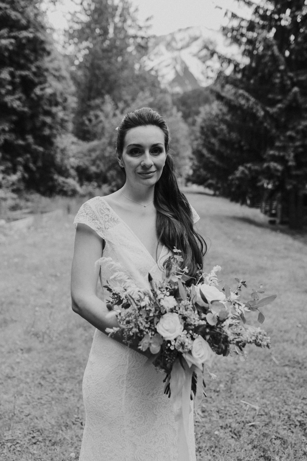 A bride in a lace wedding dress holds a beautiful wildflower bouquet against a mountain backdrop in black and white.