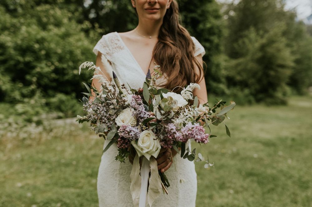 A rustic wedding bouquet with white peonies, purple wildflowers and greenery held against a lace wedding dress outdoors.