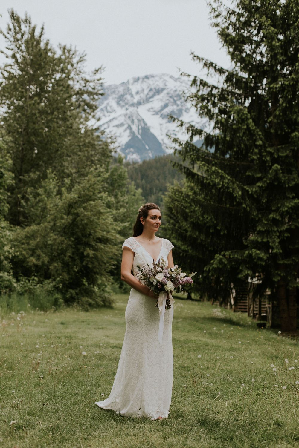 A bride in a white lace dress holds a bouquet against a backdrop of snow-capped mountains and evergreen trees.