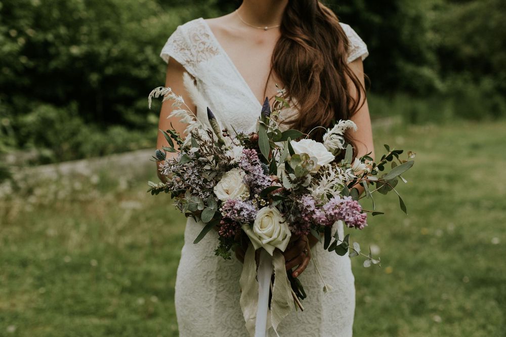 Bride holding a rustic bouquet with white and purple flowers against a green outdoor background.
