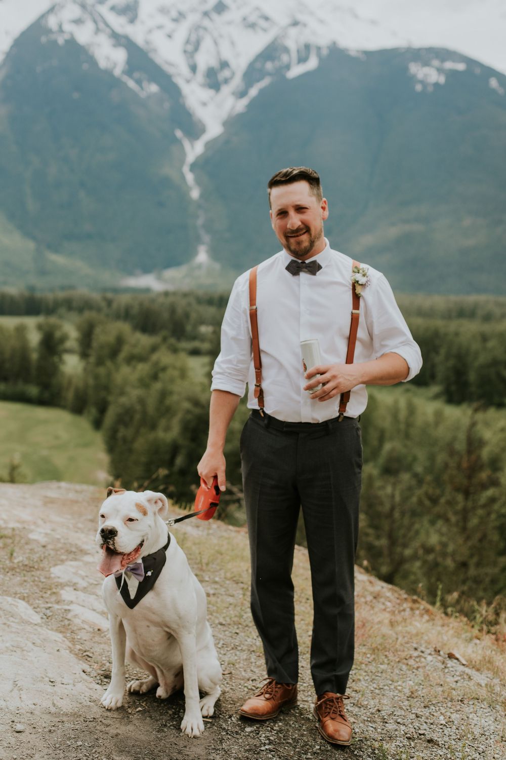 A white dog and person in suspenders pose together against a scenic mountain backdrop.