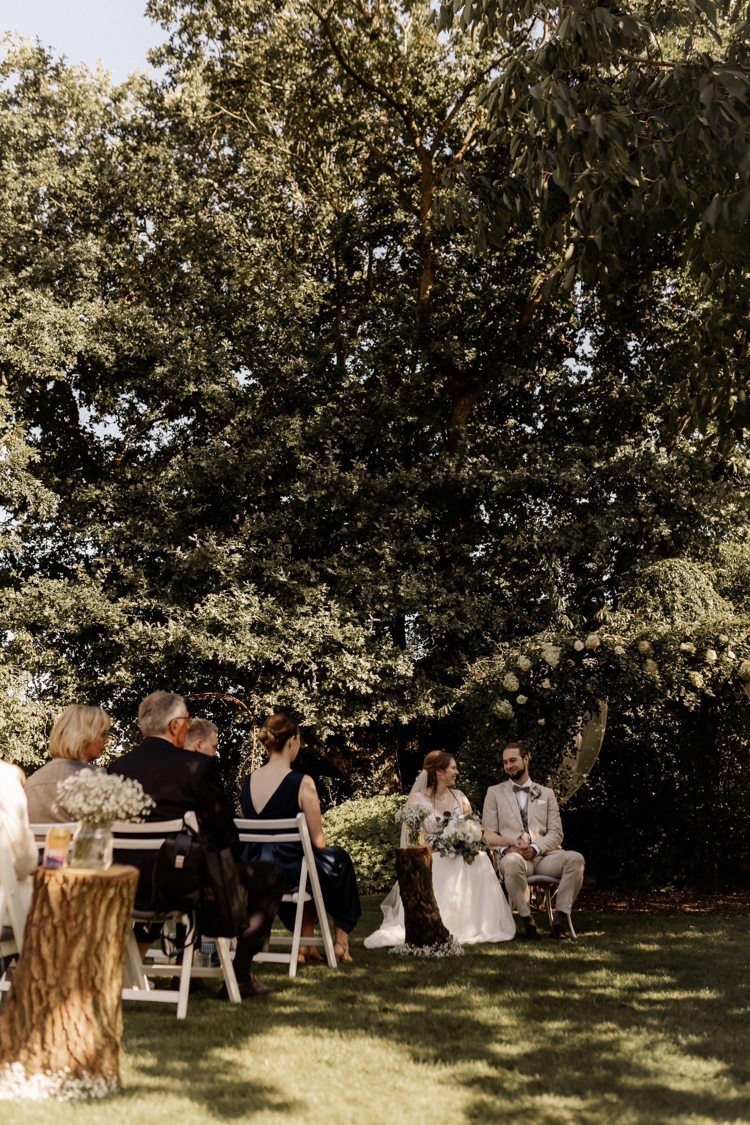 Outdoor wedding ceremony with bride and groom seated beneath a large tree surrounded by guests.