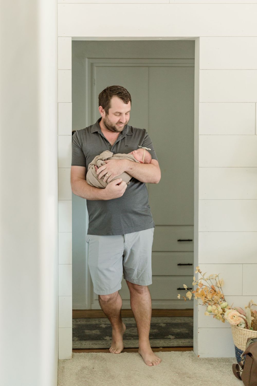 Person wearing a gray t-shirt and light shorts holds a baby while standing in a doorway with fall decor.