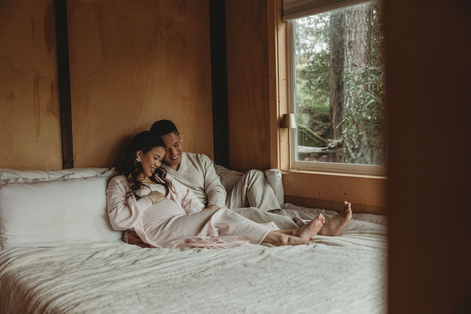 A cozy bedroom scene with a person relaxing on white bedding while gazing out a window into nature.