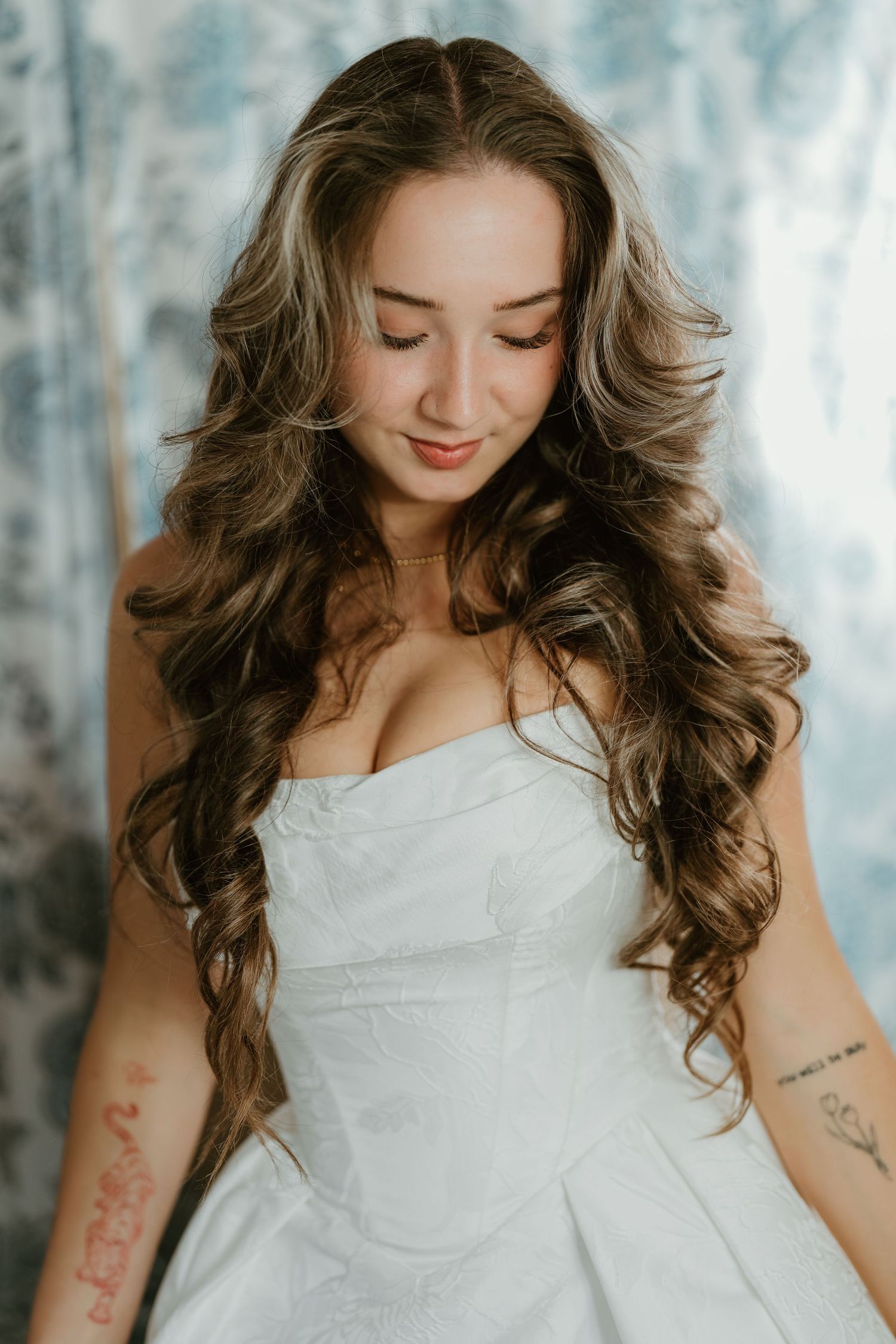 A woman in a white dress poses with flowing brown curls against a soft, ethereal background.