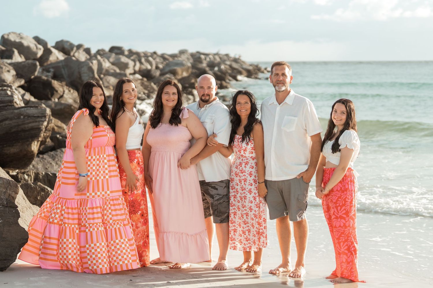 A group in coordinating coral and white outfits poses together on a sunny beach with ocean waves and rocks visible.
