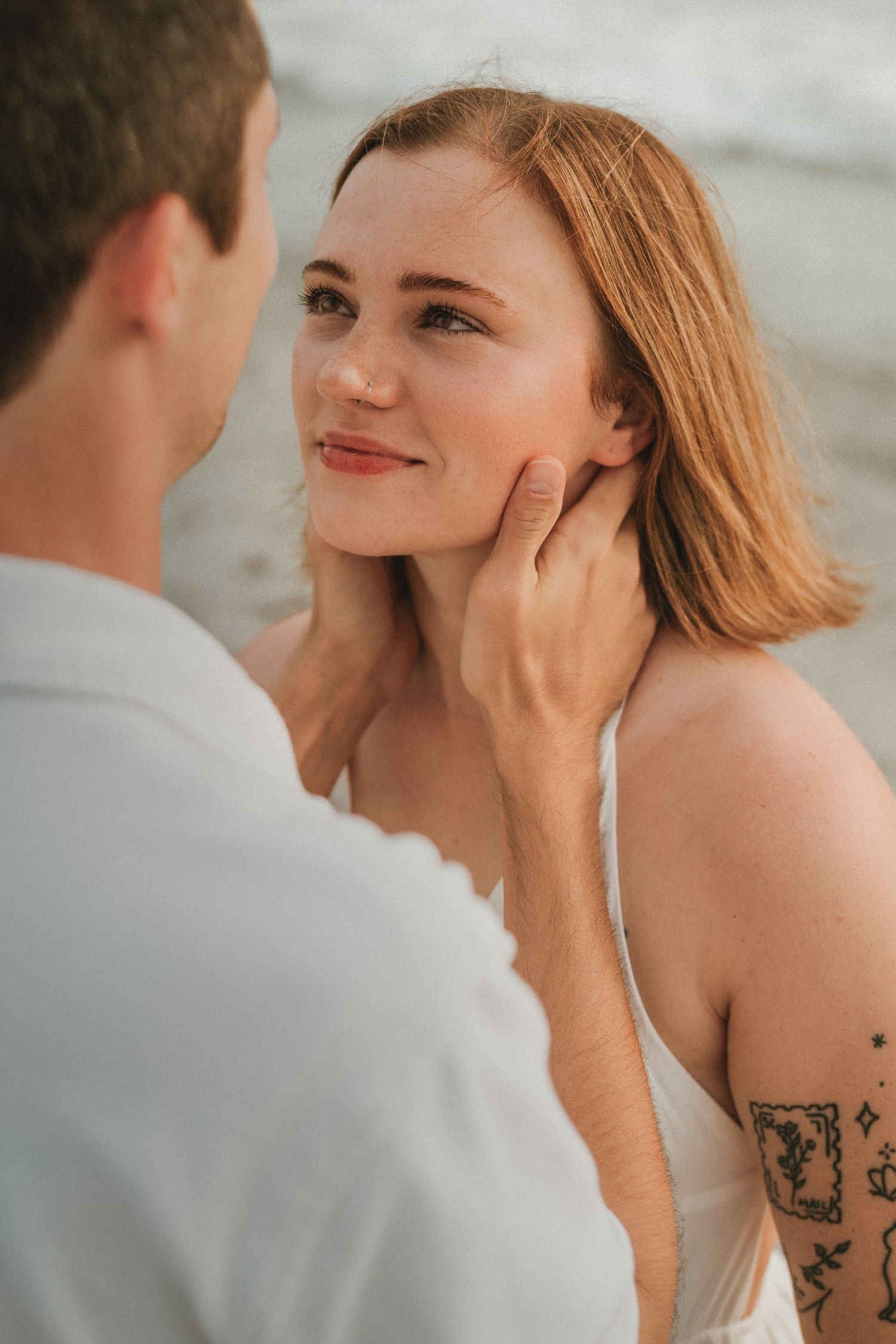 A man tenderly cups a smiling redhead woman's face on a beach, she has a tattoo on her arm.
