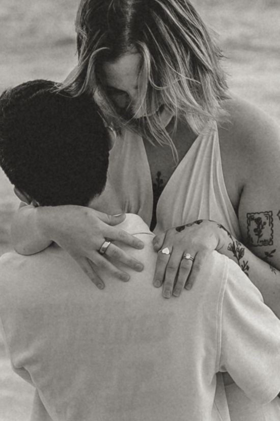Black and white photo of a couple embracing on a beach, woman with tattoos and rings leaning in close to her partner.