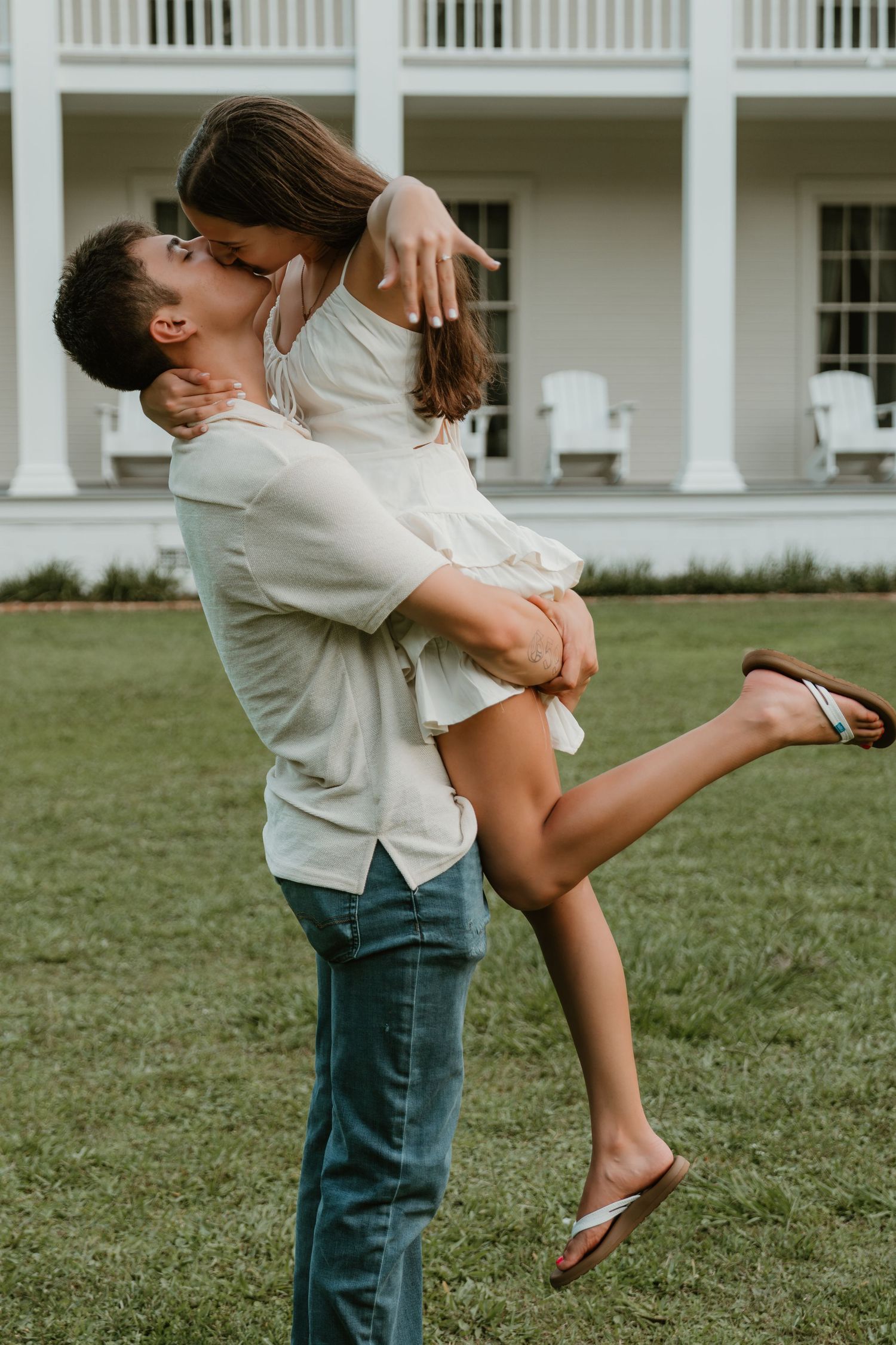 A romantic couple embraces playfully on a grassy lawn in front of a white columned building.
