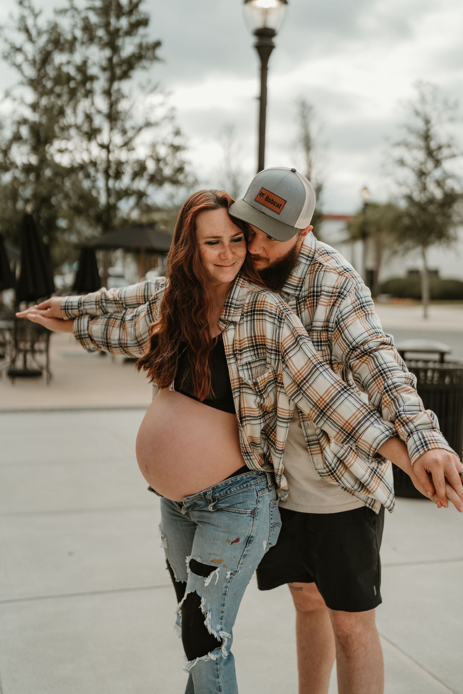 A couple in casual attire playfully dances together on a paved outdoor area during a cloudy day.