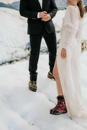 A couple in wedding attire wearing hiking boots while standing on a snowy mountain slope.