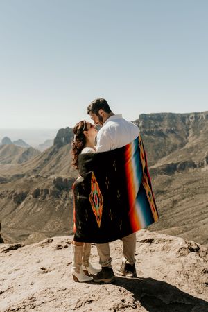 A couple wrapped in a colorful blanket stands on a mountain overlook with scenic desert views.