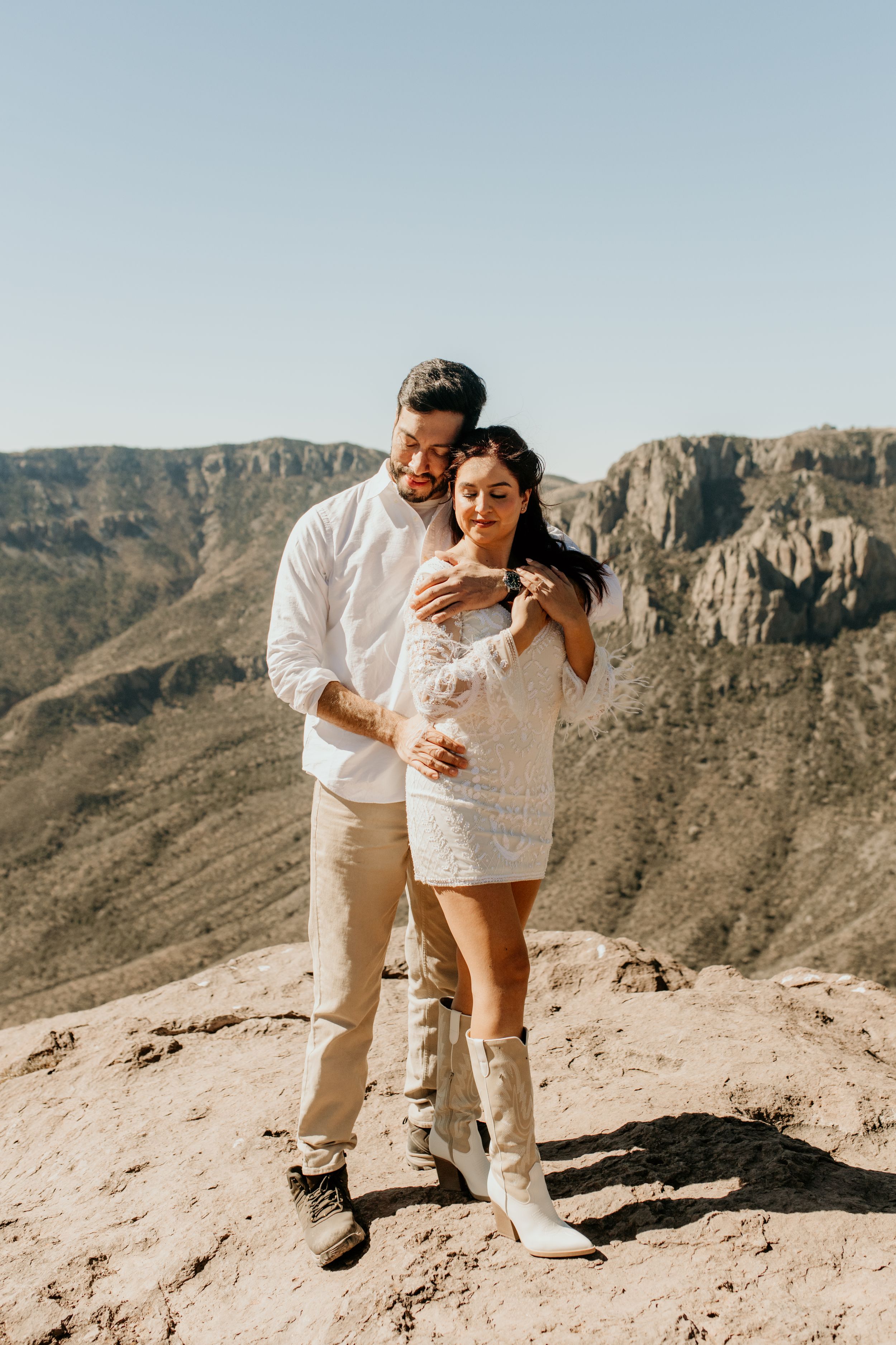 Couple in white outfits shares playful moment on desert mountain overlook.