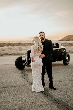 A couple poses romantically in front of a vintage hot rod car in a desert setting at sunset.