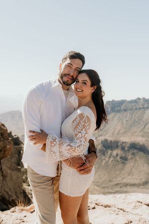 Casual beach engagement photos with couple wearing white and beige against rocky coastal backdrop.