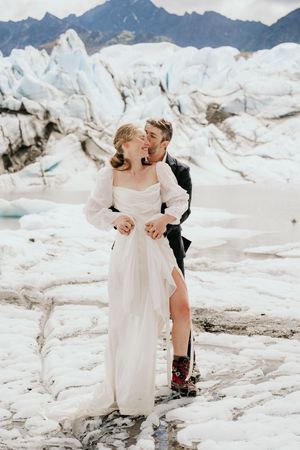 A couple in wedding attire embracing on a snowy glacier with mountains in the background.