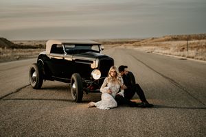 A vintage hot rod car parked on an empty desert road with a couple in wedding attire.