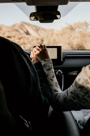 Two people holding hands in a car with mountains visible through the windshield and a GPS navigation device mounted.