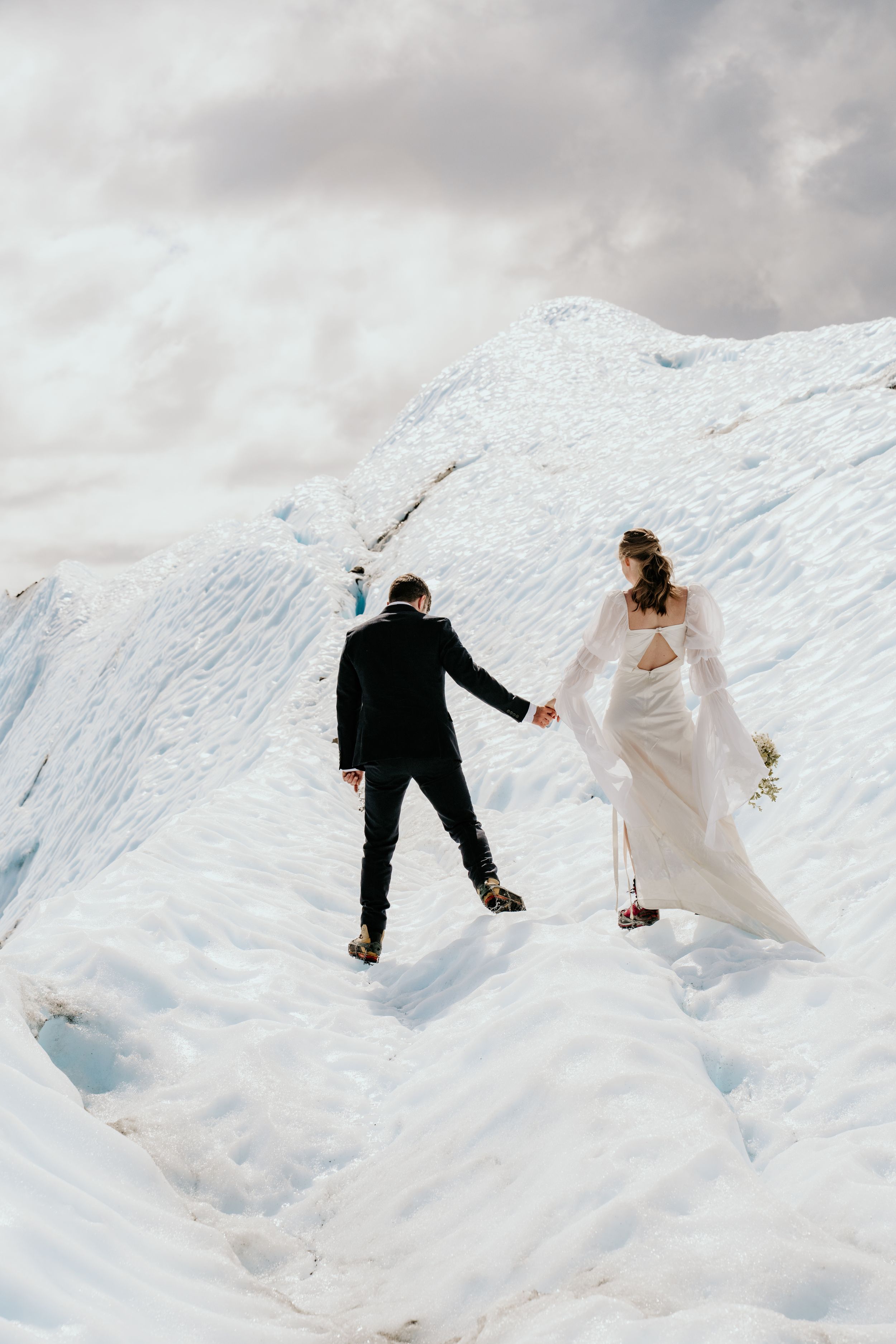 A wedding couple playfully running and sliding down a snowy slope in formal attire.