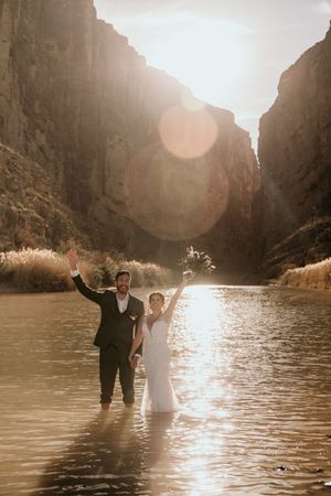 A couple celebrating in formal attire while standing in water between dramatic canyon walls at sunset.