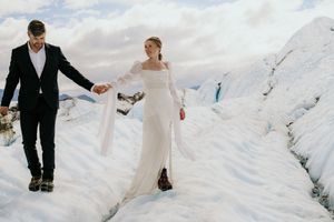 A couple in wedding attire carefully walks across a snowy glacier landscape.